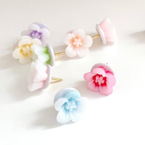 May include: Assortment of colorful flower-shaped push pins with gold heads. The pins are arranged on a white surface.