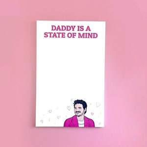 Op de afbeelding: Wit notitieblok met roze tekst die "Daddy is a state of mind" luidt. Een cartoonillustratie van een man met een snor en baard bevindt zich onderaan het notitieblok.