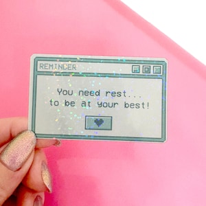 Peut inclure: Un autocollant holographique avec un design d'&eacute;cran d'ordinateur pix&eacute;lis&eacute;. L'&eacute;cran affiche le texte "REMINDER You need rest... to be at your best!" avec une petite ic&ocirc;ne de c&oelig;ur dans le coin inf&eacute;rieur droit.