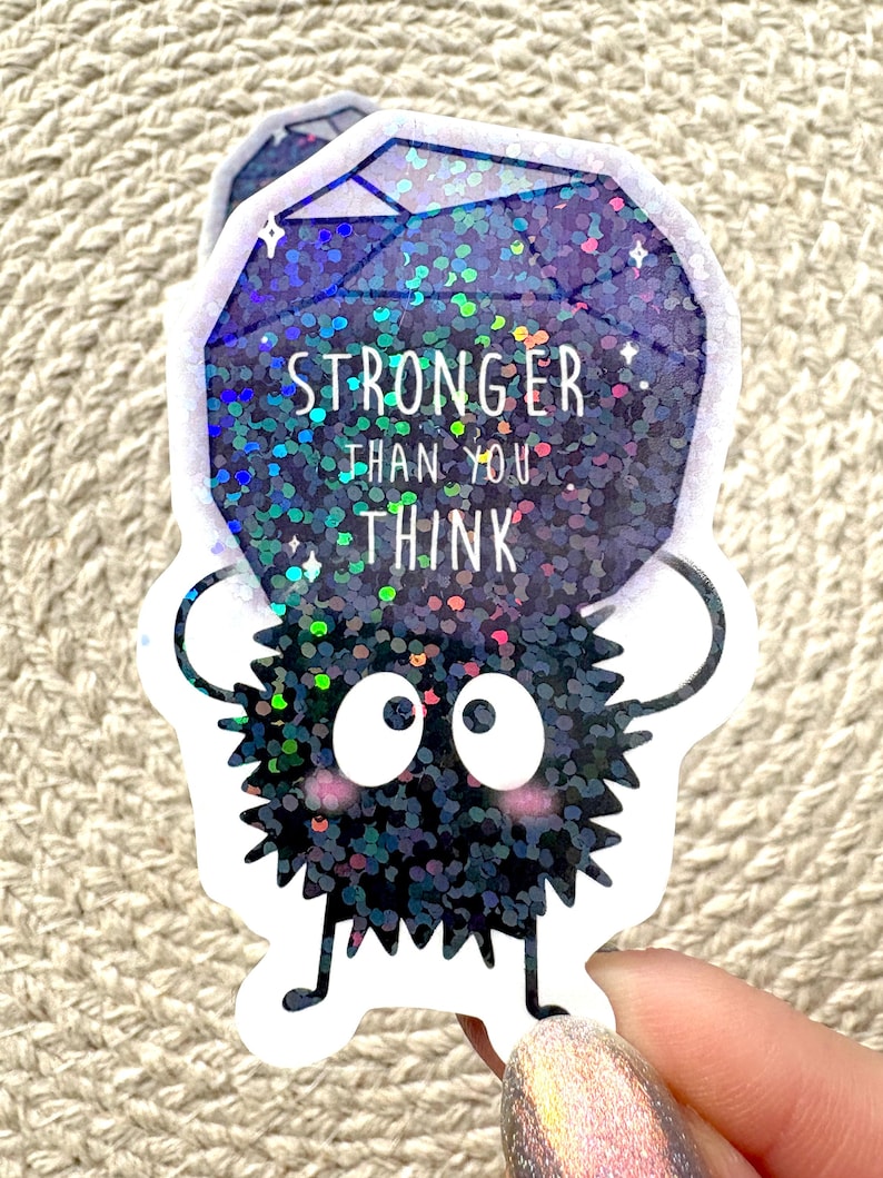 Peut inclure: Un autocollant noir et paillet&eacute; iridescent repr&eacute;sentant un personnage ressemblant &agrave; Totoro tenant un joyau violet scintillant qui dit "Stronger than you think."