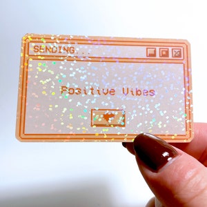 Peut inclure: Un autocollant holographique avec un fond brun clair et une finition scintillante. L'autocollant repr&eacute;sente une fen&ecirc;tre d'ordinateur avec le texte "Sending... Positive Vibes" en blanc.