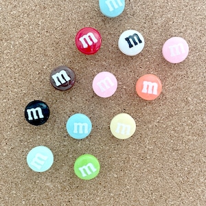 Peut inclure: Une collection de 12 punaises colorées en forme de bonbons M&M's. Les punaises sont disposées en cercle sur un tableau en liège. Les couleurs incluent le rouge, le bleu, le vert, le jaune, le rose, l'orange, le marron, le noir et le blanc.