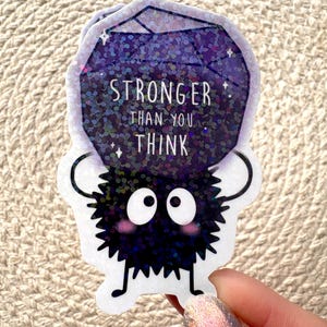 Peut inclure: Un autocollant noir et blanc d'une cr&eacute;ature floue tenant un joyau violet scintillant qui dit "Stronger than you think".