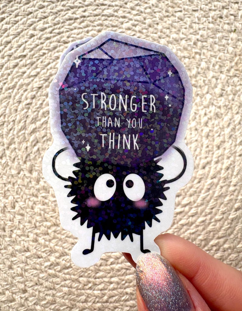 Peut inclure: Un autocollant noir et blanc d'une cr&eacute;ature floue tenant un joyau violet scintillant qui dit "Stronger than you think".