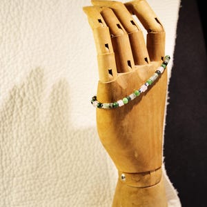 Peut inclure: Un bracelet de perles avec des pierres vertes, roses et blanches est présenté sur une main en bois. Les perles du bracelet sont petites et rondes, enfilées pour former un accessoire délicat. La main en bois est articulée et se détache sur un fond neutre.