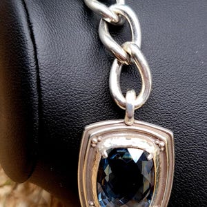 May include: Silver-toned bracelet featuring a large, square-cut, dark blue gemstone pendant. The pendant is set in a textured silver frame and connected to a chain-link bracelet. The bracelet is displayed against a black background.