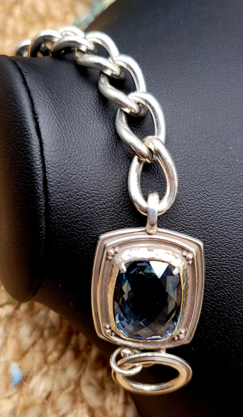 May include: Silver-toned bracelet featuring a large, square-cut, dark blue gemstone pendant. The pendant is set in a textured silver frame and connected to a chain-link bracelet. The bracelet is displayed against a black background.