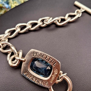 May include: Silver-toned bracelet with a large, rectangular blue gemstone. The bracelet features a chain link design and a rectangular centerpiece with the words "PEACEFUL PRESENCE" engraved around the gemstone. A toggle clasp secures the bracelet.