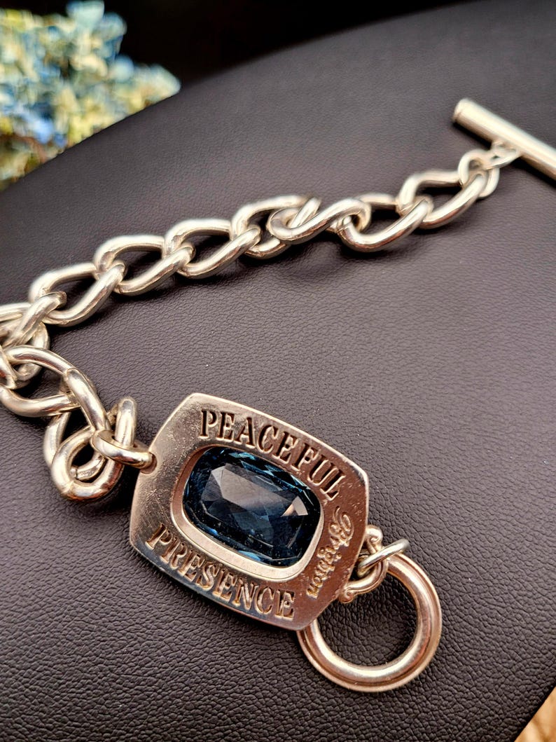 May include: Silver-toned bracelet with a large, rectangular blue gemstone. The bracelet features a chain link design and a rectangular centerpiece with the words "PEACEFUL PRESENCE" engraved around the gemstone. A toggle clasp secures the bracelet.
