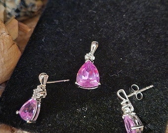 May include: A jewelry set featuring a pendant and a pair of earrings. Each piece has a pear-shaped, pink gemstone set in a silver-colored metal. The pendant and earrings have a small cluster of clear stones at the top.