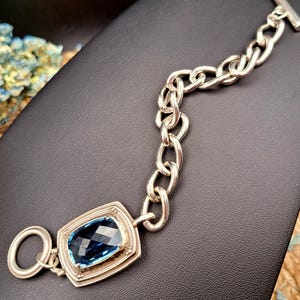 May include: Silver-toned bracelet featuring a large, rectangular, faceted blue gemstone. The bracelet has a chunky chain design and a toggle clasp. The gemstone is set in a decorative bezel. The bracelet is displayed on a black surface.