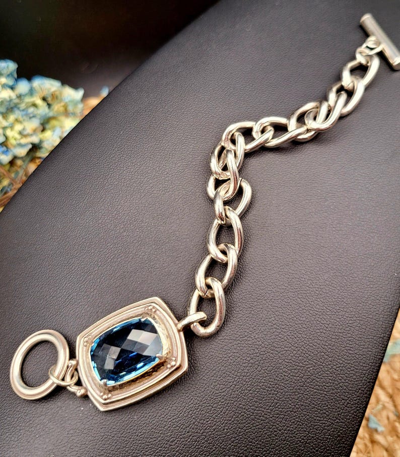 May include: Silver-toned bracelet featuring a large, rectangular, faceted blue gemstone. The bracelet has a chunky chain design and a toggle clasp. The gemstone is set in a decorative bezel. The bracelet is displayed on a black surface.