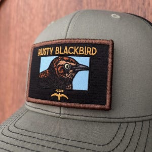 May include: A gray baseball cap with a black mesh back and a rectangular patch. The patch features the text "RUSTY BLACKBIRD" above a detailed illustration of a bird with brown and black feathers, set against a light blue background.