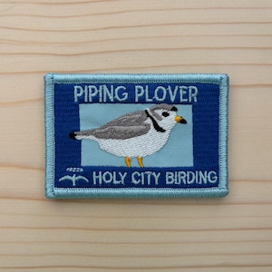 May include: Embroidered patch featuring a Piping Plover bird illustration. The patch has a blue border and a blue background with the text "PIPING PLOVER" and "HOLY CITY BIRDING". The bird is gray, white, and yellow.