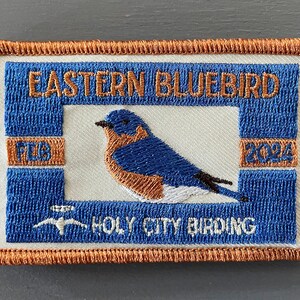 May include: An embroidered patch featuring an Eastern Bluebird with the text "Eastern Bluebird" and "Holy City Birding" in orange thread. The patch also includes the year "2024" and the letters "FLB" in orange thread.