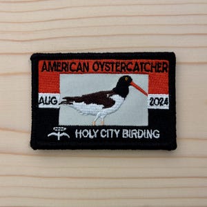 May include: Embroidered patch featuring an American Oystercatcher bird. The patch has a black border with red and white accents. Text includes "AMERICAN OYSTERCATCHER", "AUG 2024", and "HOLY CITY BIRDING". The bird is depicted with brown, white, and black feathers and a long red beak.