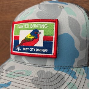 May include: A light blue and gray camouflage baseball cap with a mesh back. The cap features a rectangular patch with the words "PAINTED BUNTING" and "HOLY CITY BIRDING" surrounding a colorful bird illustration.