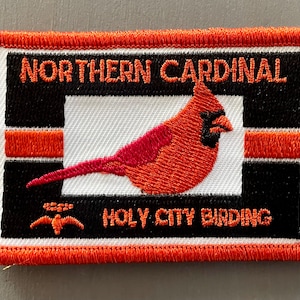 May include: An orange embroidered patch with a black border and white center. The patch features a red cardinal bird with a black beak and eye. The text "NORTHERN CARDINAL" is above the bird and "HOLY CITY BIRDING" is below the bird.
