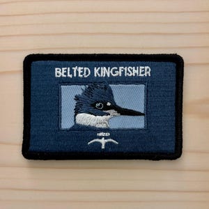 May include: A rectangular embroidered patch with a dark blue border and background. It features a light blue center panel with an embroidered head of a belted kingfisher bird, the text BELTED KINGFISHER above, and a small white bird icon below.