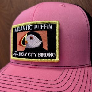 May include: Pink trucker hat with a black mesh back. The front features a rectangular patch with the words "ATLANTIC PUFFIN" above an illustration of a puffin bird and the words "HOLY CITY BIRDING".