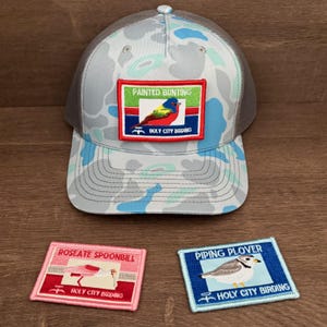 May include: A camouflage-patterned trucker hat with a gray mesh back and a patch featuring a Painted Bunting bird. Two embroidered patches below display a Roseate Spoonbill and a Piping Plover, both with "Holy City Birding" text.