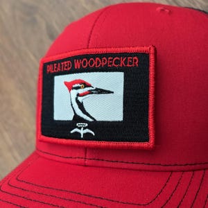 May include: Red baseball cap featuring a rectangular patch with the text "PILEATED WOODPECKER" above an embroidered image of a woodpecker. The woodpecker is depicted in black, white, and red. The cap has a curved brim.