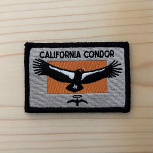 May include: A rectangular embroidered patch with a black border. The background is light gray with an orange center rectangle. It features a black California Condor with white wing patches and the text CALIFORNIA CONDOR in black above the bird.