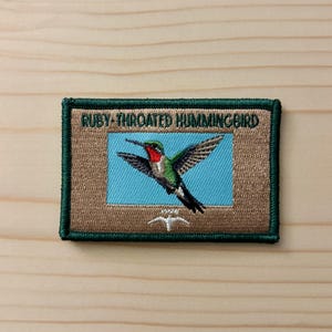 May include: Embroidered patch featuring a Ruby-Throated Hummingbird with outstretched wings. The patch has a brown border with the text "RUBY-THROATED HUMMINGBIRD" above the bird. The hummingbird is green, red, and gray, set against a blue background.