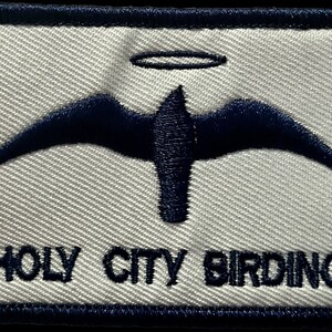 May include: Embroidered patch with a navy blue bird design and a halo above it. The patch has the words "HOLY CITY BIRDING" embroidered in navy blue. The patch is rectangular with a navy blue border and a light gray background.