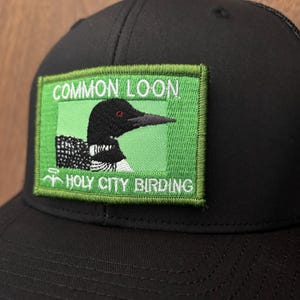 May include: Black baseball cap with a green patch featuring a Common Loon illustration. The patch includes the text "COMMON LOON" and "HOLY CITY BIRDING". The bird is black and white with a red eye.