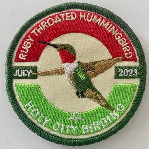 May include: A green, red, and white embroidered patch with a hummingbird in flight. The patch reads "Rusty Throated Hummingbird" and "Holy City Birding" with the dates "July" and "2023".