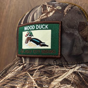 May include: A camouflage baseball cap with a patch featuring a wood duck illustration. The patch has the text "WOOD DUCK" above the duck and "HOLY CITY BIRDING" below. The cap has a brown mesh back.