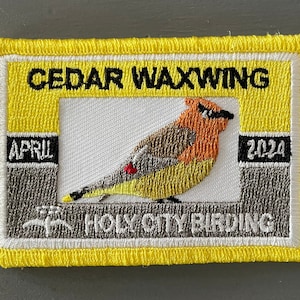 May include: A yellow embroidered patch with a cedar waxwing bird illustration. The patch says "CEDAR WAXWING" at the top, "APRIL 2024" on the sides, and "HOLY CITY BIRDING" at the bottom.