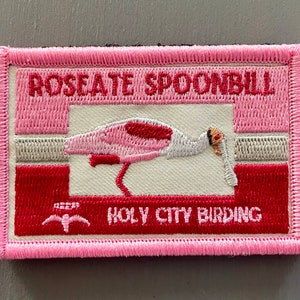 May include: Embroidered patch featuring a Roseate Spoonbill bird in shades of pink and red, with the text "ROSEATE SPOONBILL" and "HOLY CITY BIRDING". The patch has a pink border and a rectangular shape, suitable for bird enthusiasts.