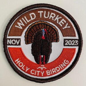 May include: A brown and orange embroidered patch with a white background. The patch features a wild turkey with a brown body and a red head. The text "WILD TURKEY" is in brown embroidery around the top of the patch. The text "NOV 2023" is in white embroidery on the left and right sides of the patch. The text "HOLLY CITY BIRDING" is in white embroidery around the bottom of the patch.