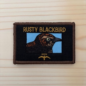 May include: A rectangular embroidered patch with a brown border and a black background. It features the text RUSTY BLACKBIRD in gold thread above a light blue square containing an embroidered profile of a dark bird with a sharp beak.