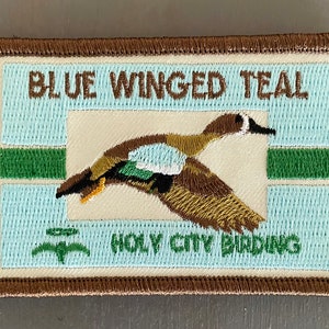 May include: A brown embroidered patch with a blue-winged teal flying in front of a white background with green stripes. The text "BLUE WINGED TEAL" is above the bird and "HOLY CITY BIRDING" is below the bird.