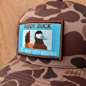 May include: A brown and tan camouflage baseball cap with a rectangular patch featuring a ruddy duck illustration. The patch has the text "RUDDY DUCK" and "HOLY CITY BIRDING" in white lettering on a blue background.