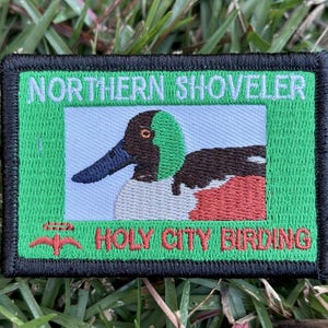 May include: Embroidered patch featuring a Northern Shoveler bird illustration. The patch has a green border with the text "NORTHERN SHOVELER" and "HOLY CITY BIRDING" in white and red. The bird has a blue beak, brown head, and red and white chest.