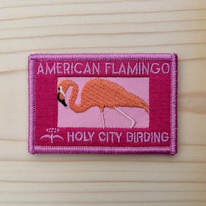 May include: Embroidered patch featuring an orange flamingo on a pink background, with the text "AMERICAN FLAMINGO" above and "HOLY CITY BIRDING" below. The patch has a pink border and is set against a light wood grain.