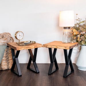 May include: Two wooden side tables with live edges and black metal X-shaped legs. One table has a gold clock and tray, the other has a lamp with a glass base and white shade. The tables are on a dark wood floor.