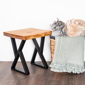 May include: A small, square wooden table with a natural wood top and black metal X-shaped legs. A woven basket filled with blankets and throws sits nearby. The table is approximately 18 inches long.