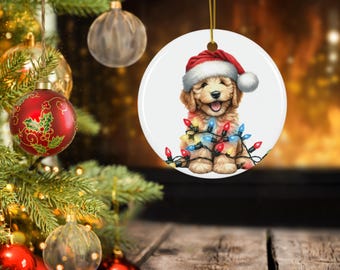 May include: A circular Christmas tree decoration showing a smiling puppy wearing a Santa hat and wrapped in colourful Christmas lights. The ornament has a gold string for hanging. A Christmas tree and fireplace are in the background.