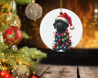May include: A circular Christmas tree decoration showing a black dog wearing a Santa hat and wrapped in multi-coloured fairy lights. The ornament hangs from a gold string. A Christmas tree and other baubles are in the background.
