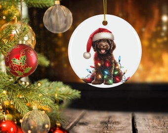 May include: A circular Christmas decoration depicting a brown dog in a Santa hat, adorned with colourful fairy lights. The ornament is suspended by a gold string, with a Christmas tree and other baubles visible in the background.