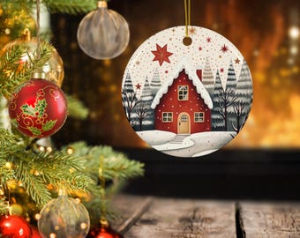 May include: A circular Christmas tree decoration depicting a red house with a snowy roof and trees. The ornament has a gold hanging string and a winter scene design. Other baubles and a Christmas tree are in the background.