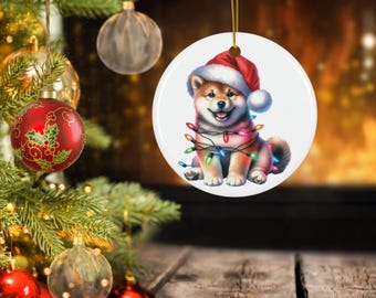 Op de afbeelding: Een ronde kerstversiering met een Shiba Inu-puppy met een kerstmuts en omwikkeld met kleurrijke kerstverlichting. De versiering is wit met een gouden touwtje om op te hangen. De achtergrond toont een kerstboom en een open haard.