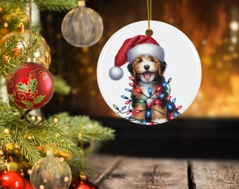 May include: A round Christmas ornament featuring a puppy wearing a Santa hat and wrapped in colorful Christmas lights. The ornament is hanging from a gold string, with a Christmas tree and other ornaments in the background.