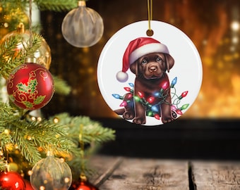 Puede incluir: Adorno navideño redondo con un cachorro de Labrador marrón con un gorro de Papá Noel y envuelto en luces navideñas de colores. El adorno tiene una cuerda dorada para colgar. El fondo borroso muestra un árbol de Navidad y una chimenea.