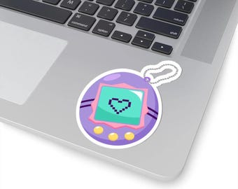 May include: A sticker featuring a retro purple virtual pet toy design. The sticker has a teal screen displaying a pixelated heart, with pink and yellow accents. The sticker is on a laptop.